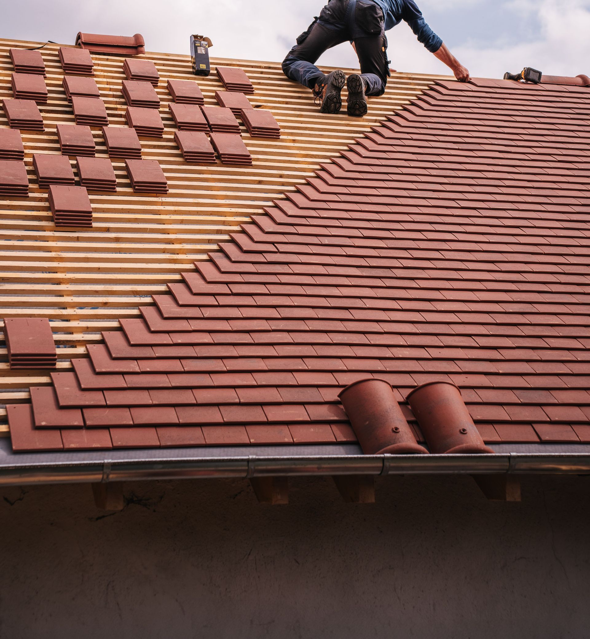 A roofer carefully installs shingles on a new residential roof under a cloudy sky.